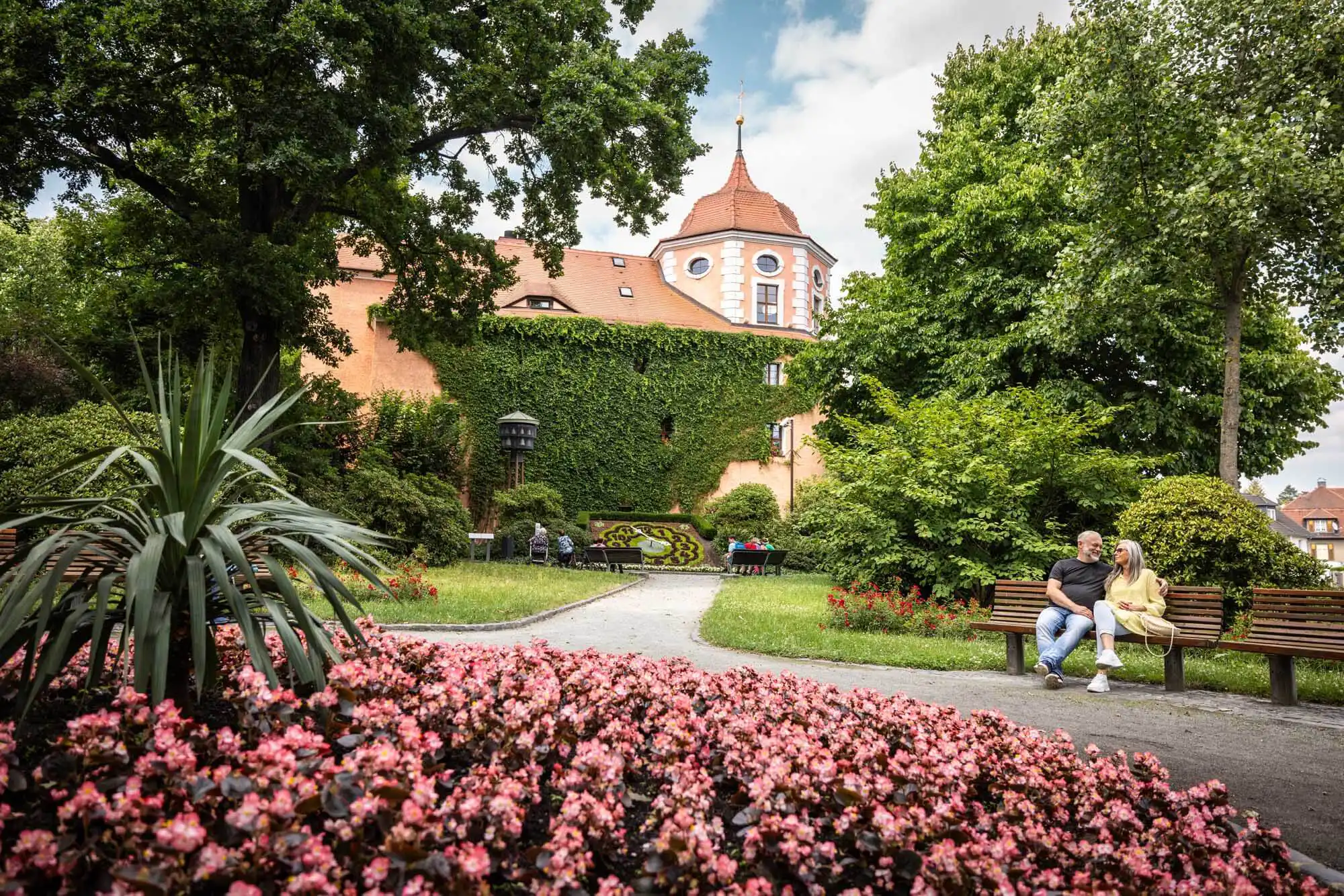 Park mit Blumenbeet und Pärchen auf Parkbank. Im Hintergrund ein historisches Gebäude